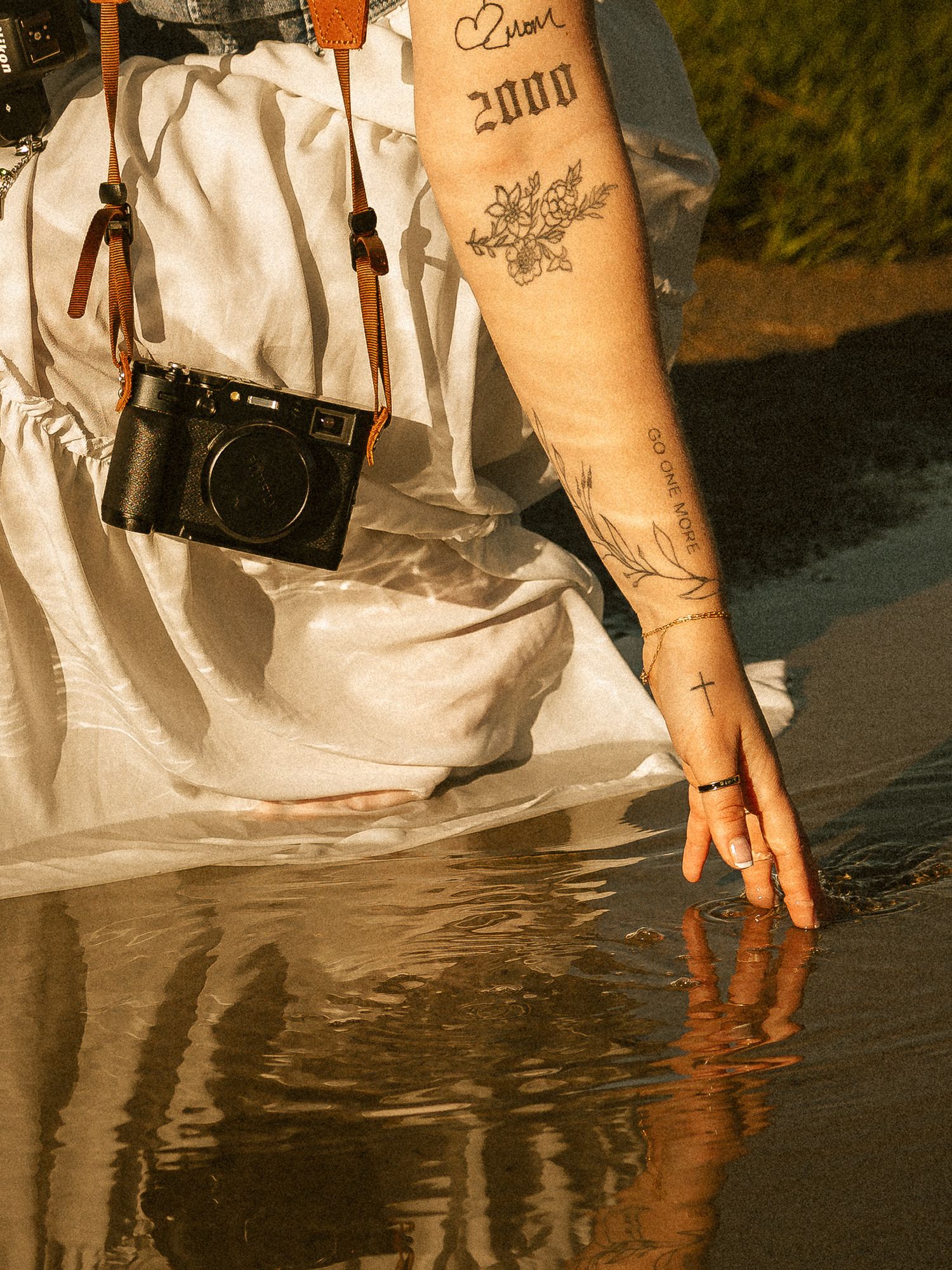 Tattooed arm reaching into water, white dress, black camera on strap, golden hour light.