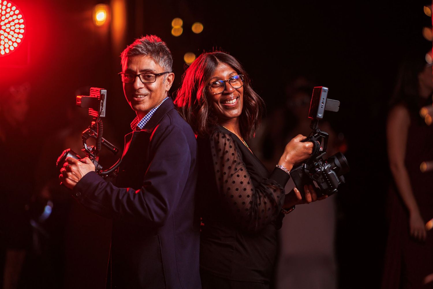 Two photographers smiling back-to-back holding cameras with lights at an evening event.