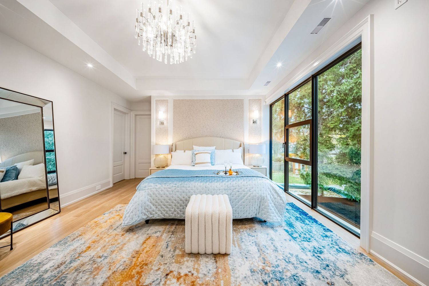 Spacious master bedroom with floor-to-ceiling windows, crystal chandelier, and blue patterned area rug.
