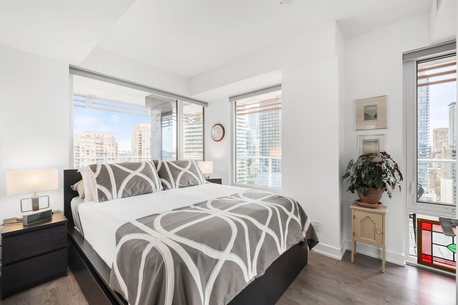 Modern bedroom with gray geometric bedding, white walls, city views through large windows, and wood flooring.