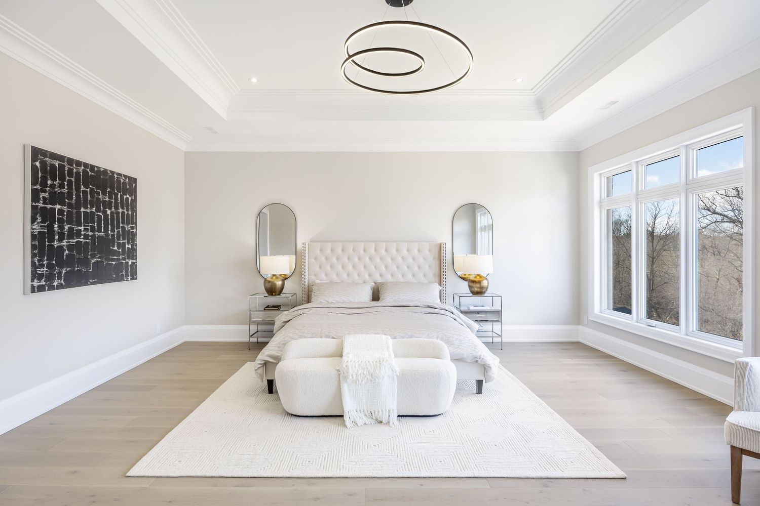 Modern white bedroom with tufted headboard, circular pendant light, and large windows offering natural illumination.