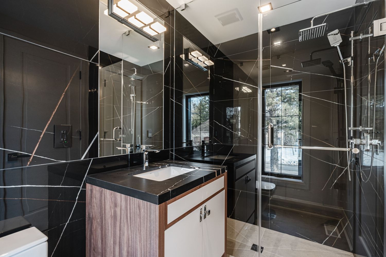 Dark modern bathroom with glass shower enclosure, marble-look tiles, and wood vanity accents.