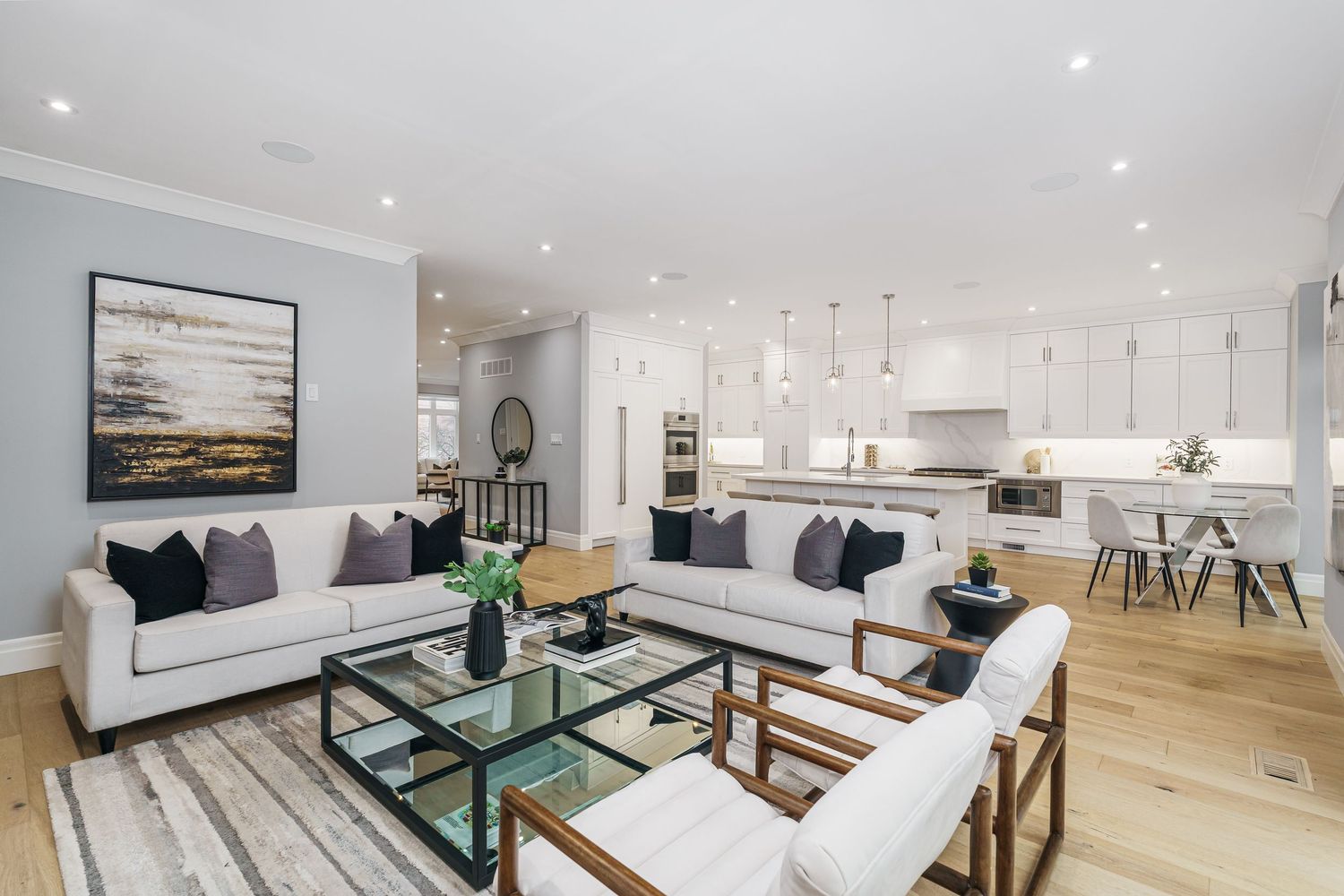 Modern open concept living room with white sofas, glass coffee table and connected kitchen featuring hardwood floors.