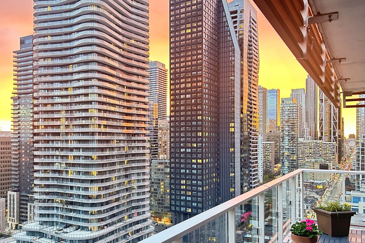 Modern high-rise apartment balcony overlooking glass skyscrapers against a dramatic orange sunset in downtown Toronto.