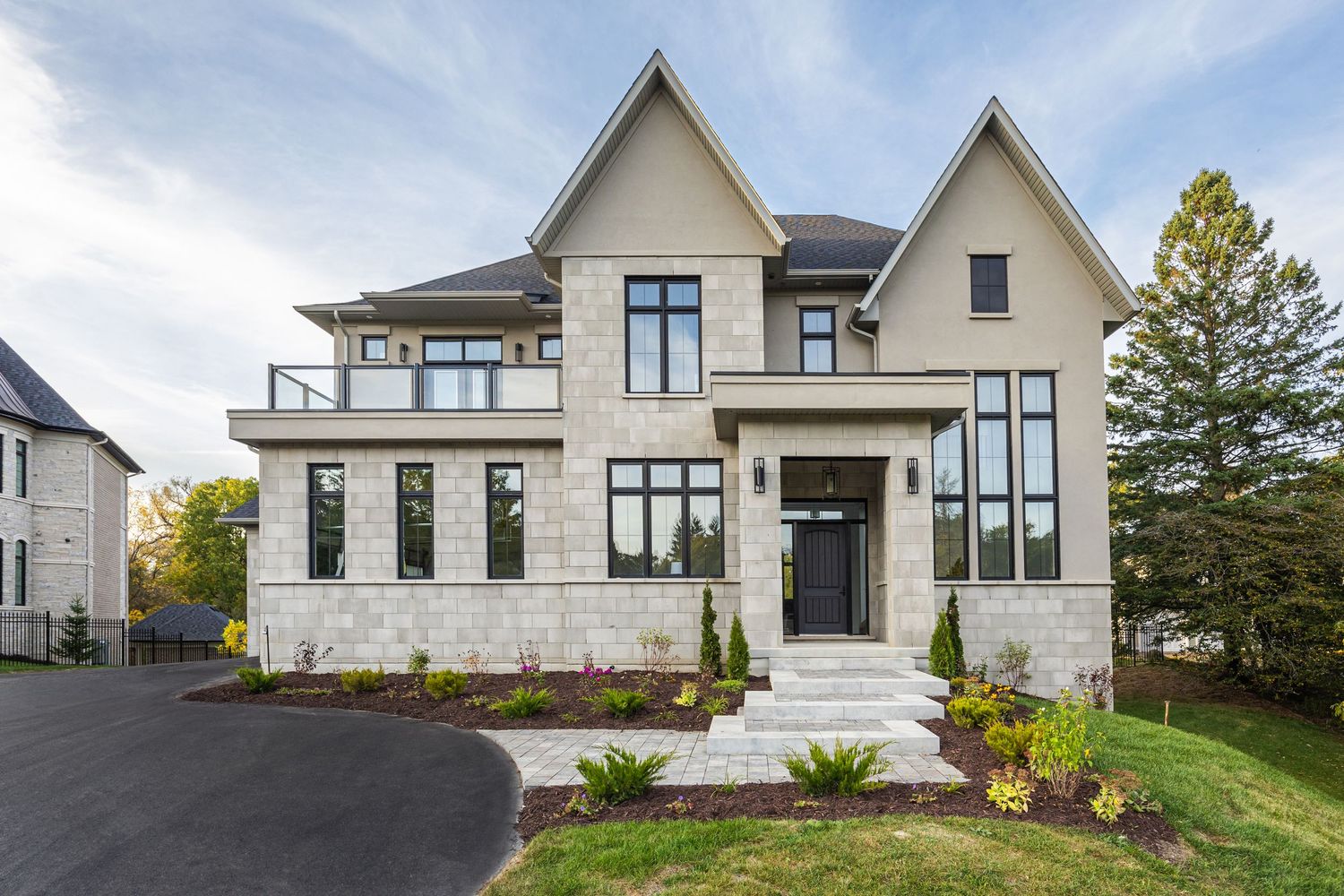 Contemporary white stone mansion with large windows, double peaked roof, and landscaped front yard.
