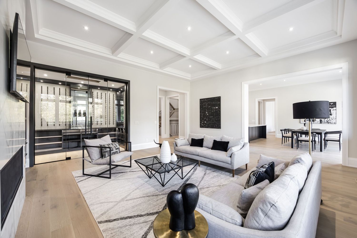 Modern luxury living room with white coffered ceiling, hardwood floors, and glass-enclosed wine display.