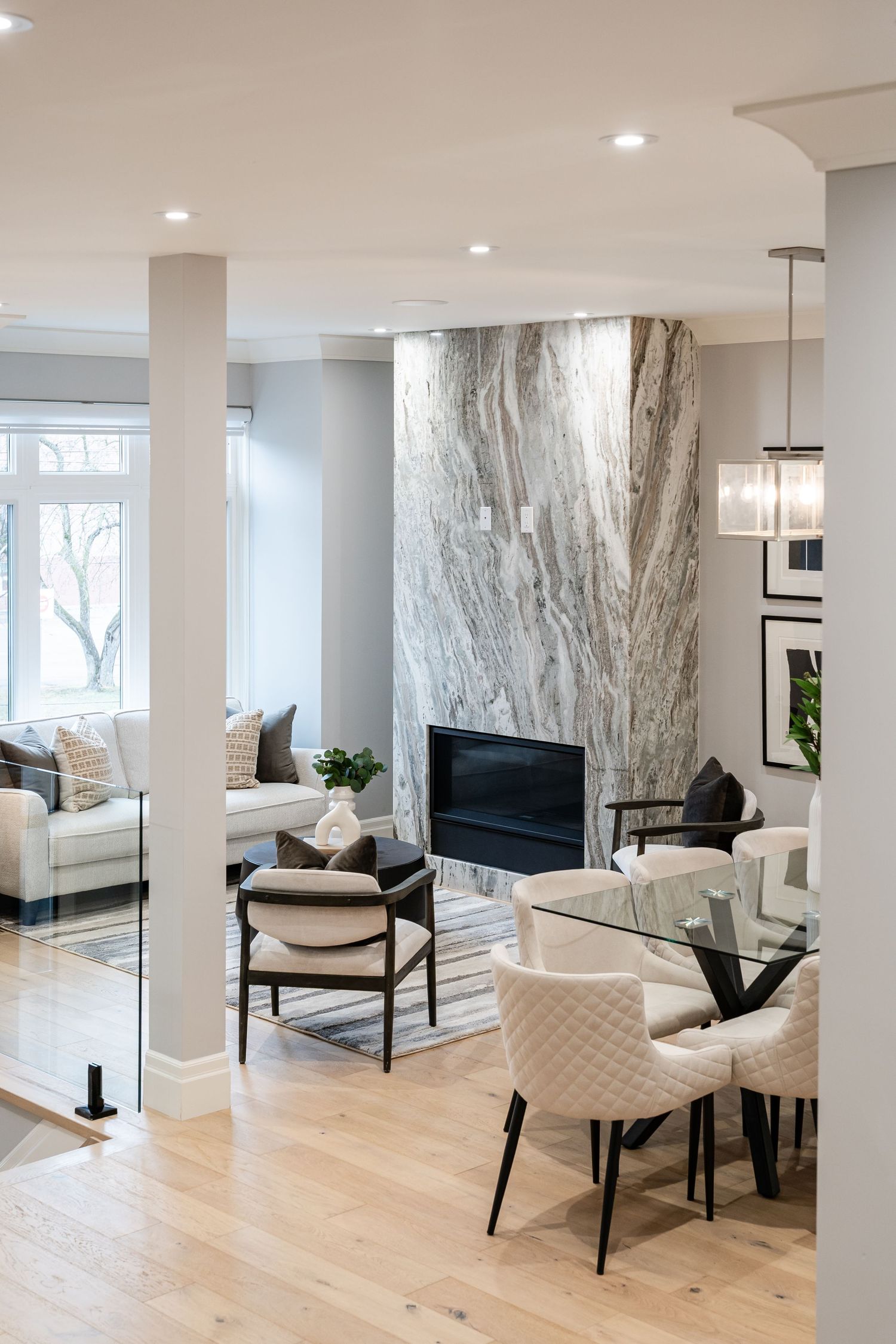 Modern living room with marble fireplace wall, light wood floors, white chairs and natural lighting from large windows.