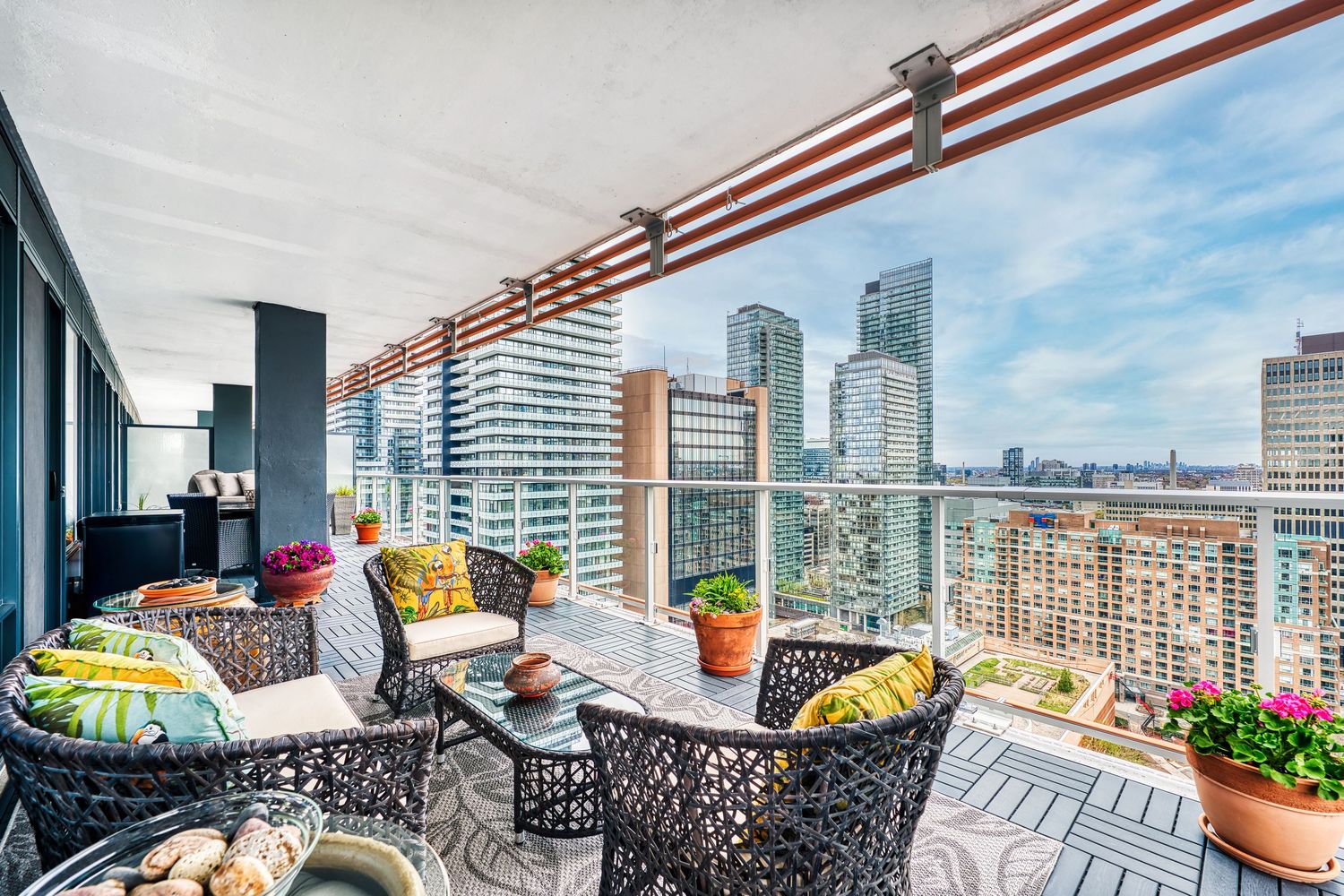 Modern balcony with woven chairs and yellow pillows overlooking city skyline with high-rise buildings on a sunny day.