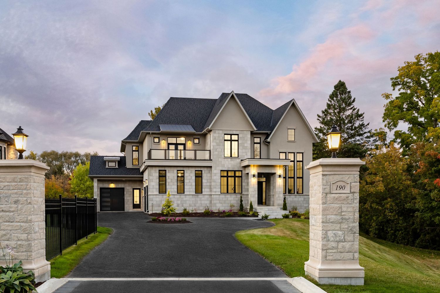 Luxurious modern mansion with grey exterior and white trim illuminated at dusk with paved driveway entrance.