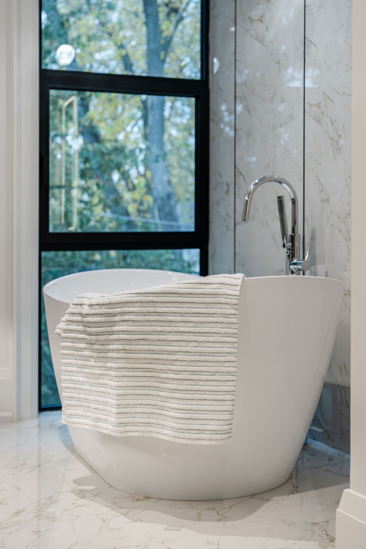 Freestanding white soaking tub with striped towel near floor-to-ceiling windows overlooking nature.