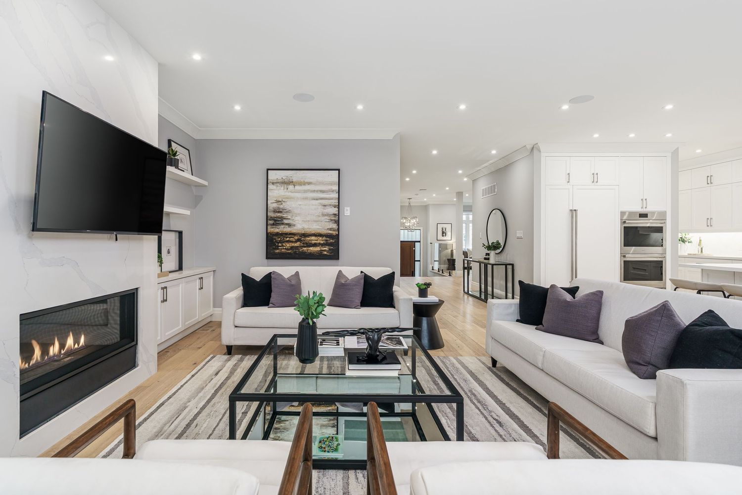Modern living room with white sofas, linear fireplace, wall-mounted TV, and glass coffee tables in a bright open concept space.