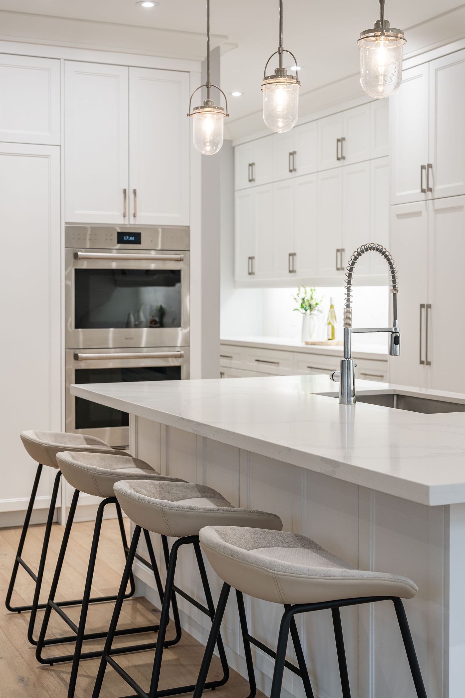Modern white kitchen with pendant lights, built-in stainless appliances, and gray upholstered bar stools at quartz island counter.