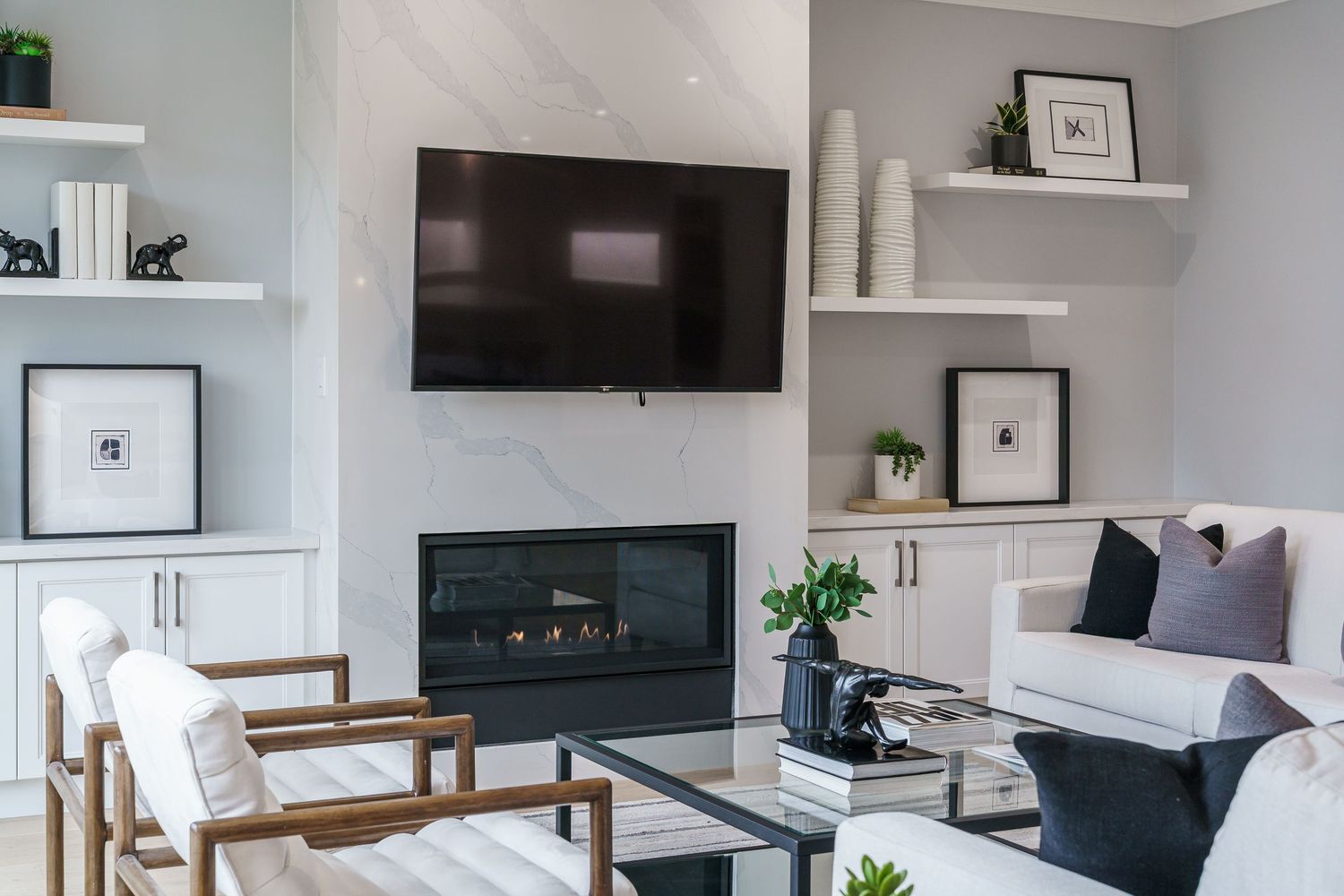 Modern living room with wall-mounted TV, fireplace, white floating shelves, and contemporary wood accent chairs.