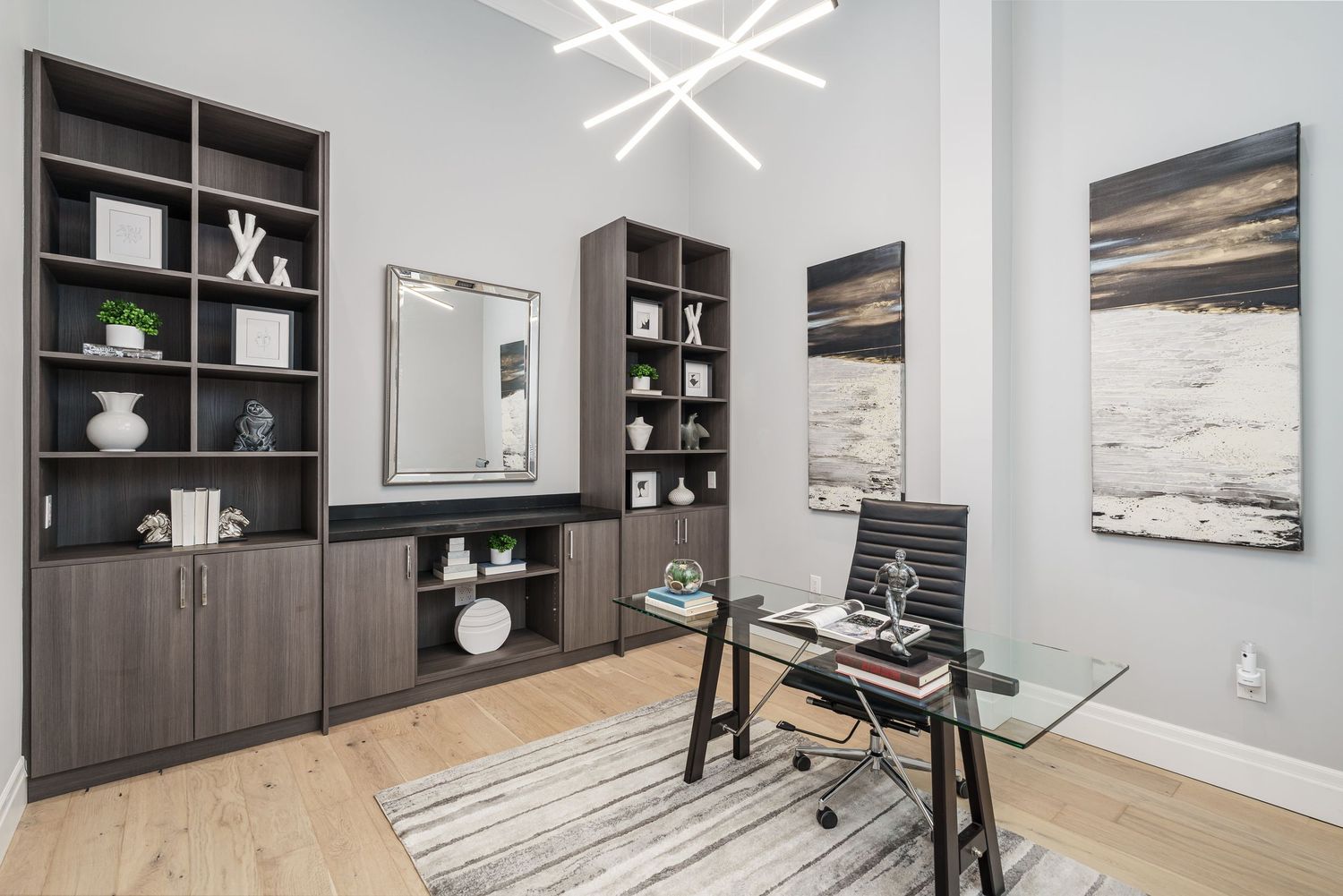 Modern home office with dark wood built-ins, glass desk, geometric light fixture and abstract wall art on hardwood floors.