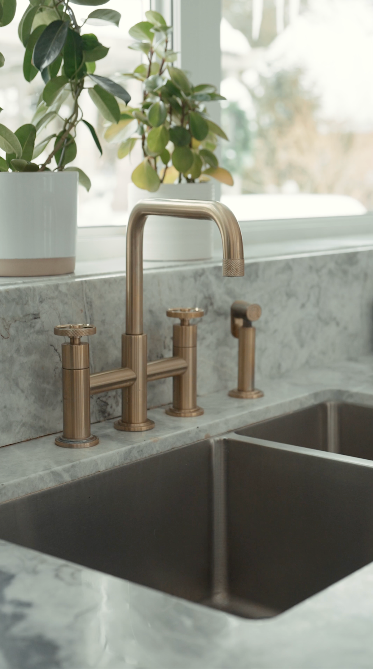 Modern brushed brass kitchen faucet and double sink with side sprayer on white marble countertop near window with plants.