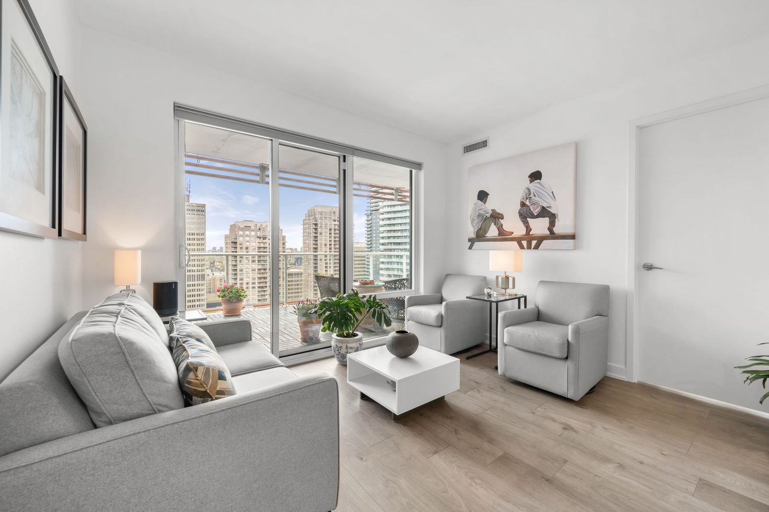 Modern living room with floor-to-ceiling windows offering city views, featuring gray furniture and hardwood floors.