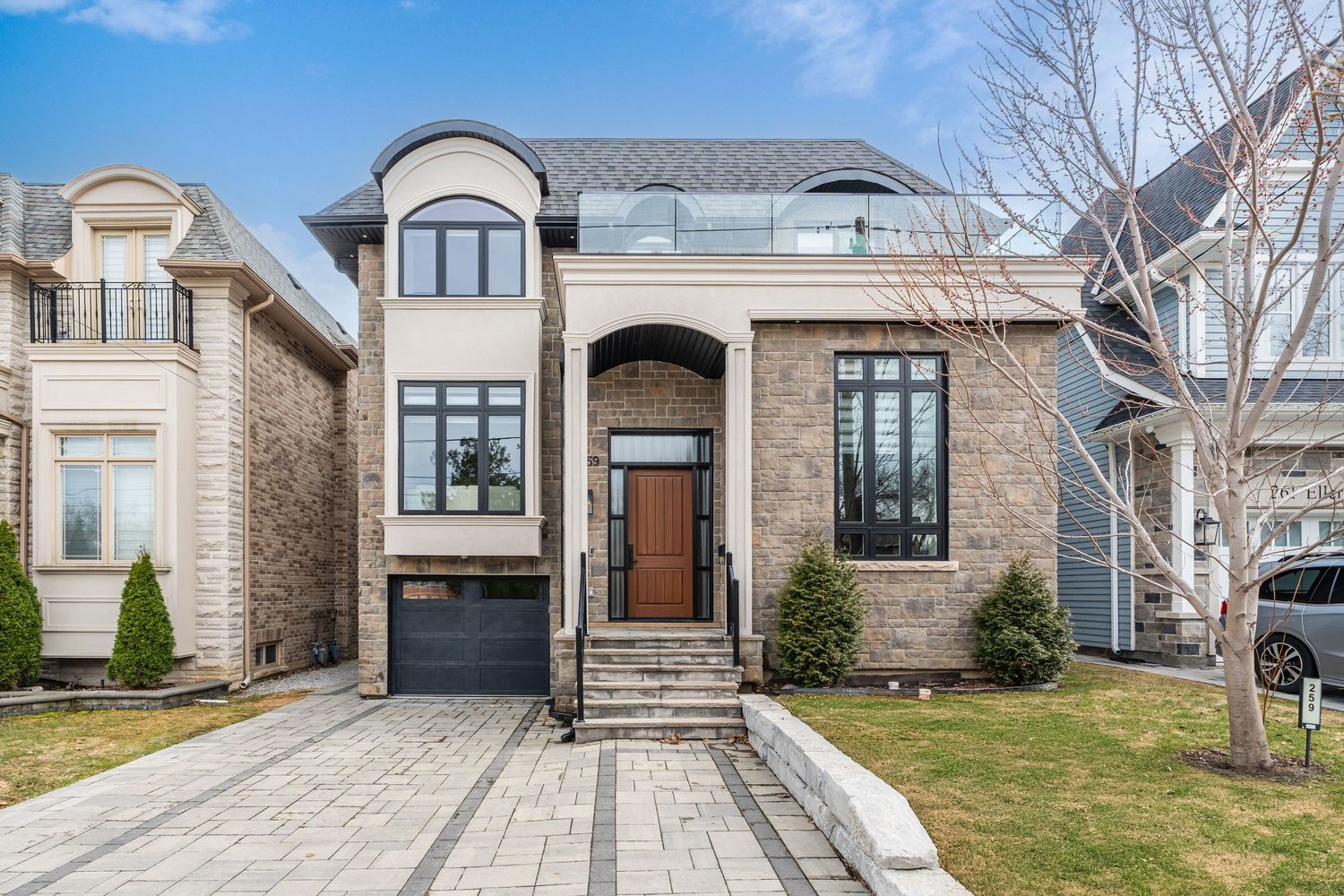 Modern stone-faced luxury home with arched windows, dark wood door and interlocking stone driveway in upscale neighborhood.