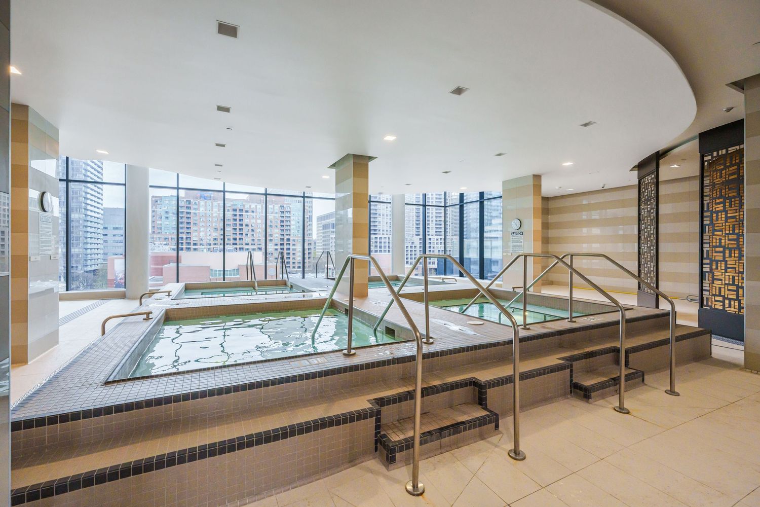 Modern indoor spa facility with large whirlpool tub, metal handrails, and floor-to-ceiling windows overlooking city buildings.