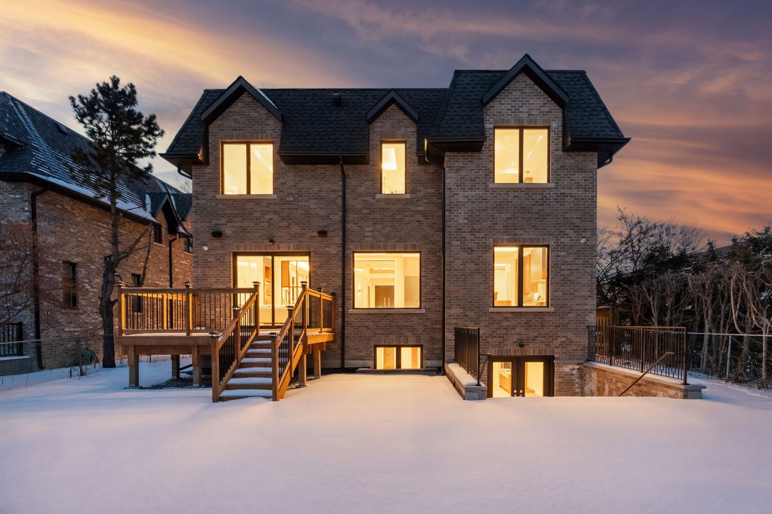 Modern stone house with illuminated windows and wooden deck photographed at sunset in snowy conditions.