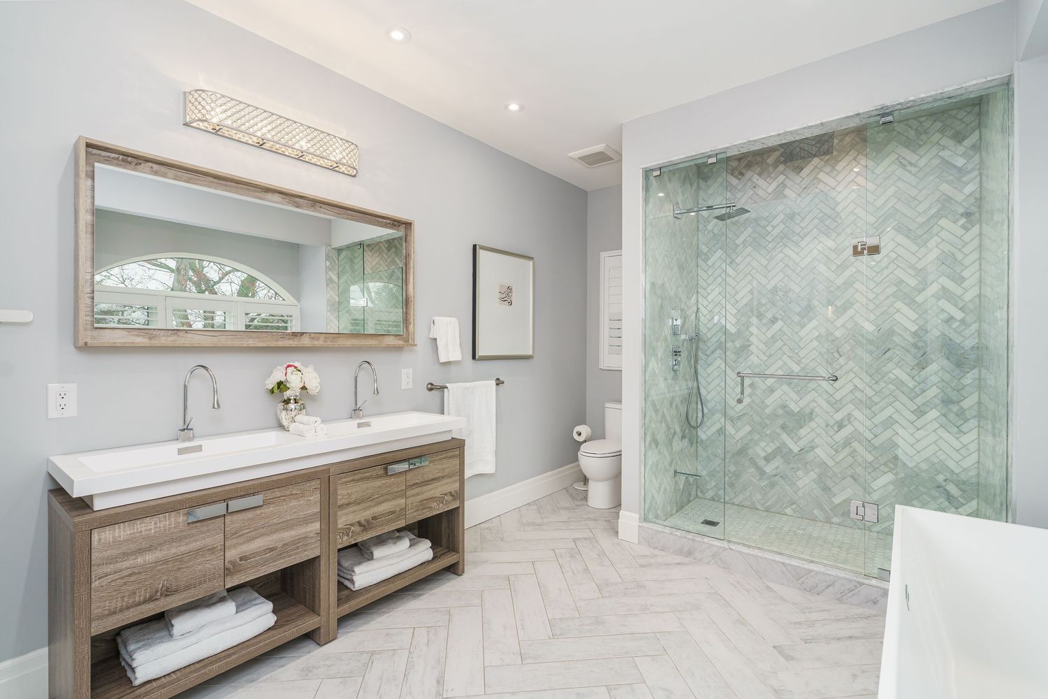 A modern master bathroom renovation showcases herringbone tile, a freestanding tub, and natural wood vanity with glass accents.