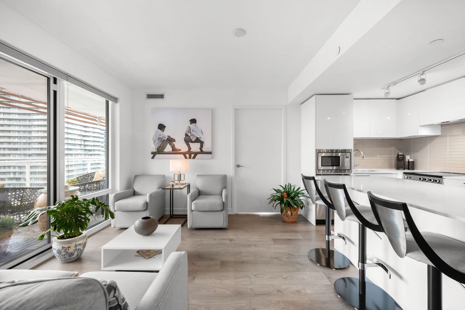 Modern open concept condo with hardwood floors, gray furniture, and white kitchen featuring large windows and plants.