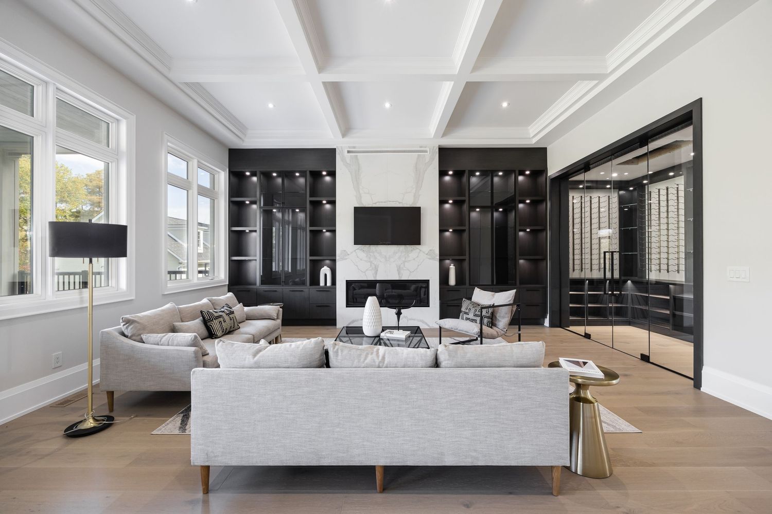 Modern luxury living room with light gray sofas, hardwood floors, coffered ceiling and black built-in shelving.