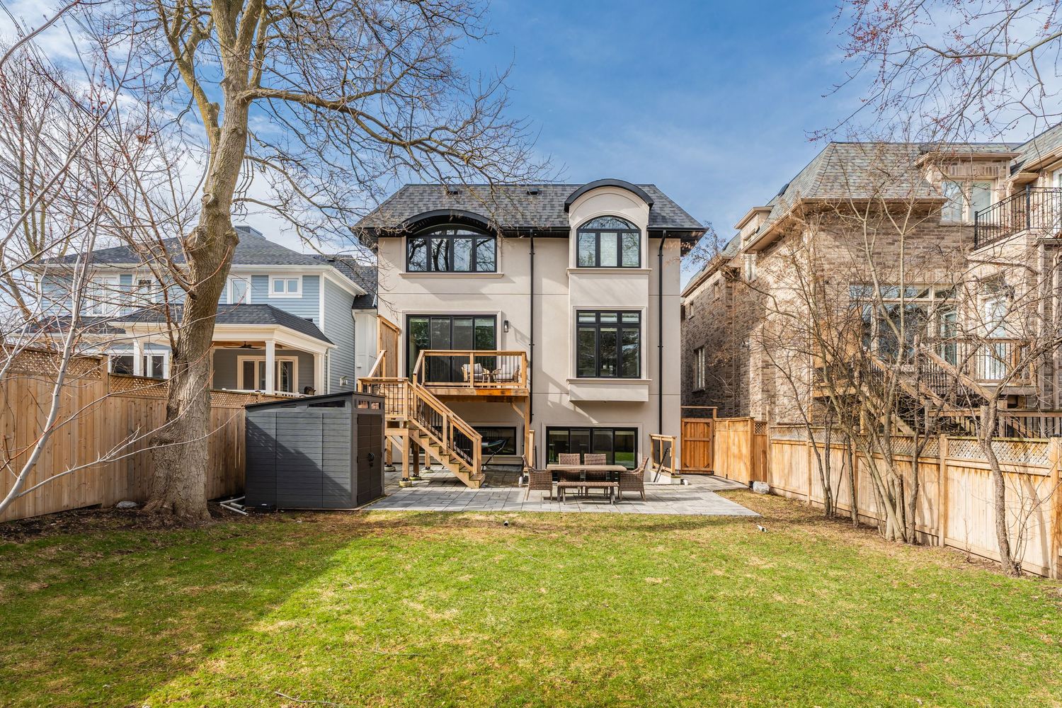 Modern three-story grey house with dormer windows and wooden deck overlooking spacious backyard on sunny day.