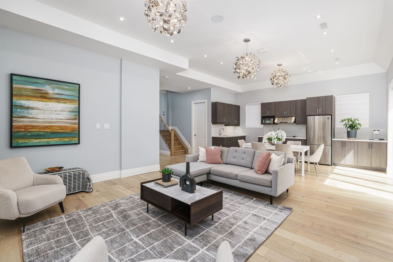 Modern open concept living room with light wood floors, grey area rug, crystal chandeliers and connected kitchen space.