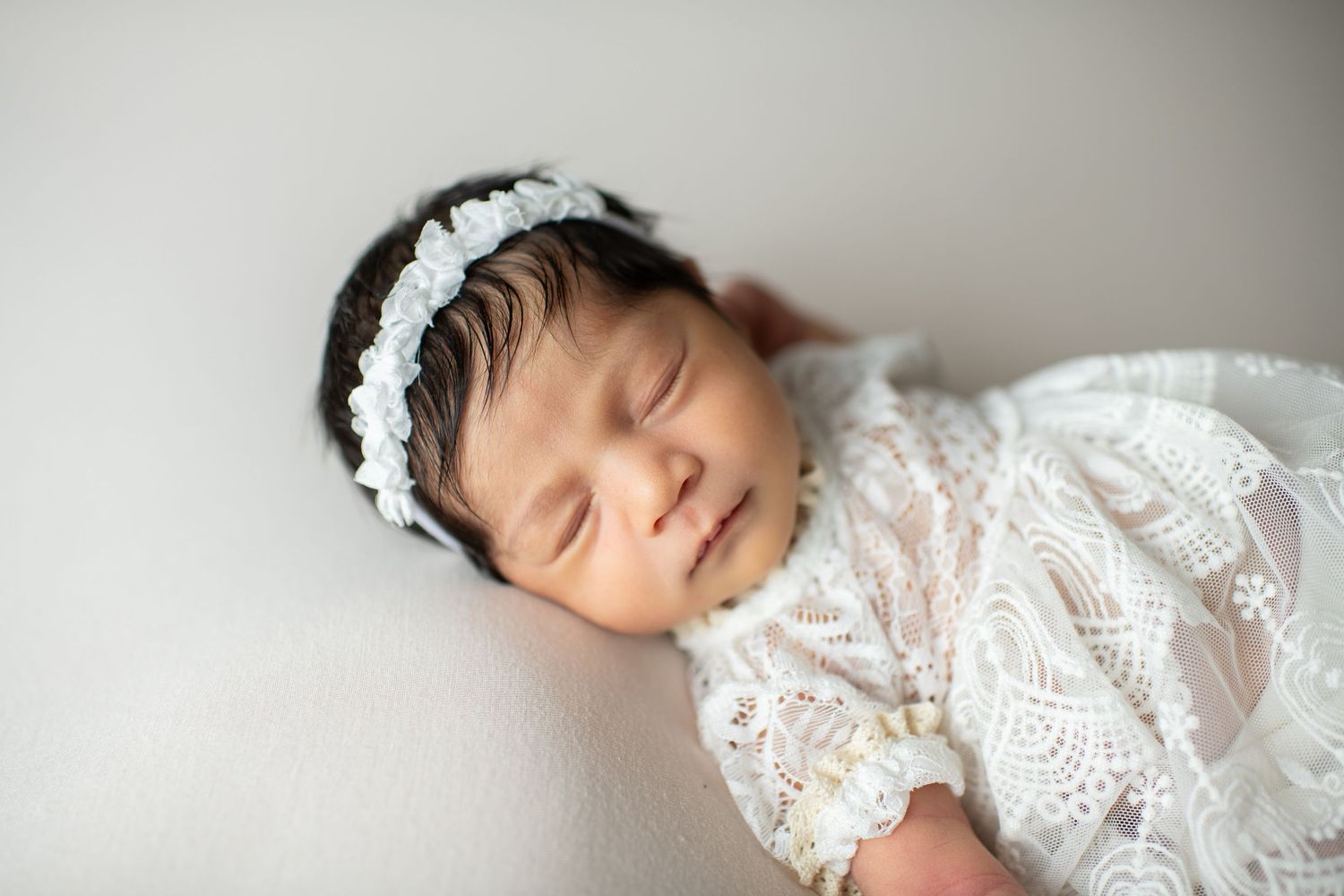 Sleeping newborn wearing delicate white lace dress and floral headband rests peacefully on light backdrop.