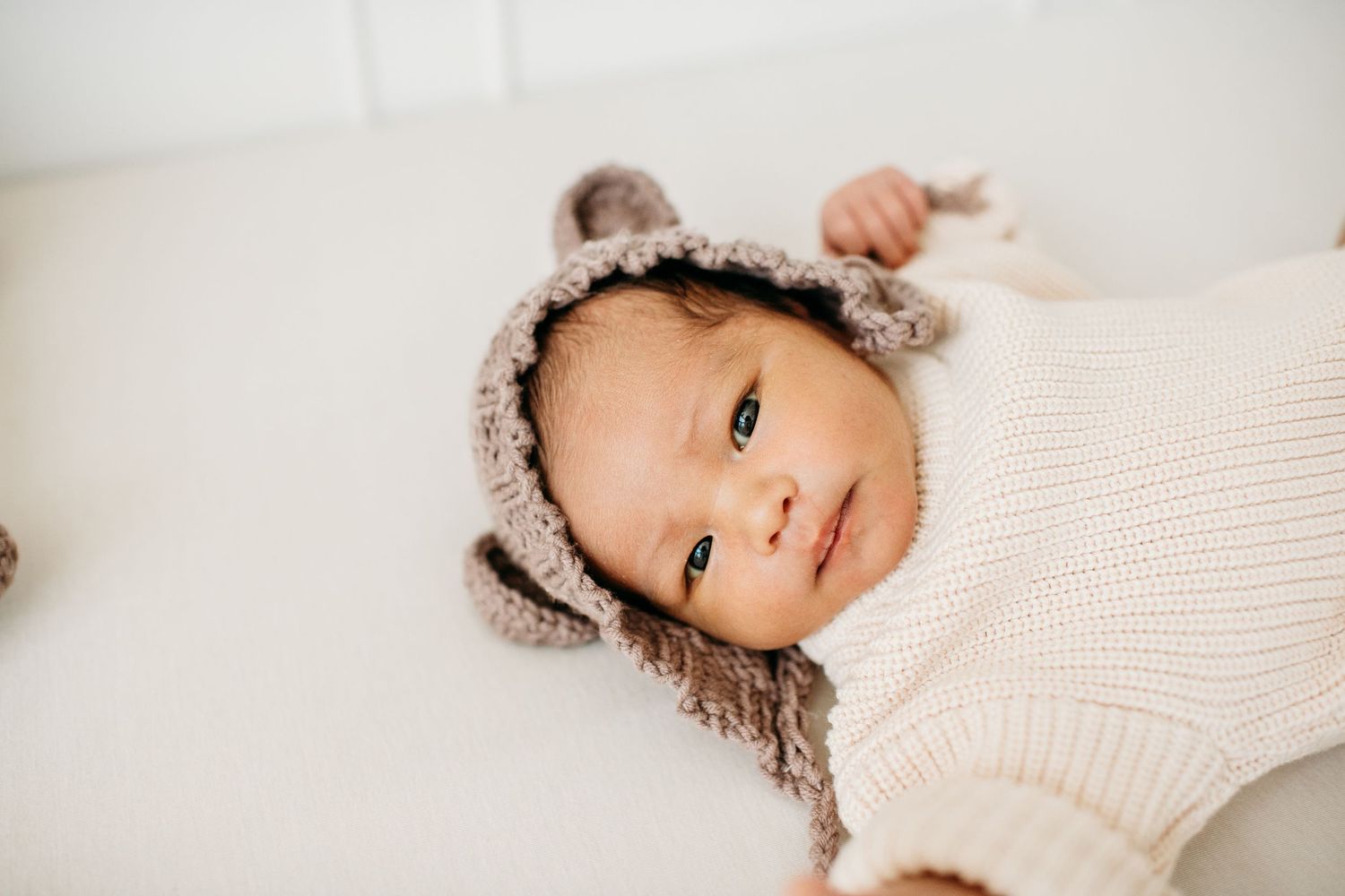 A baby wearing a cozy cream knit sweater and furry bonnet lies on white bedding.