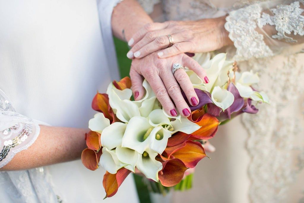 Wedding bouquet of orange and white calla lilies with hands displaying rings and lace sleeves in the frame.