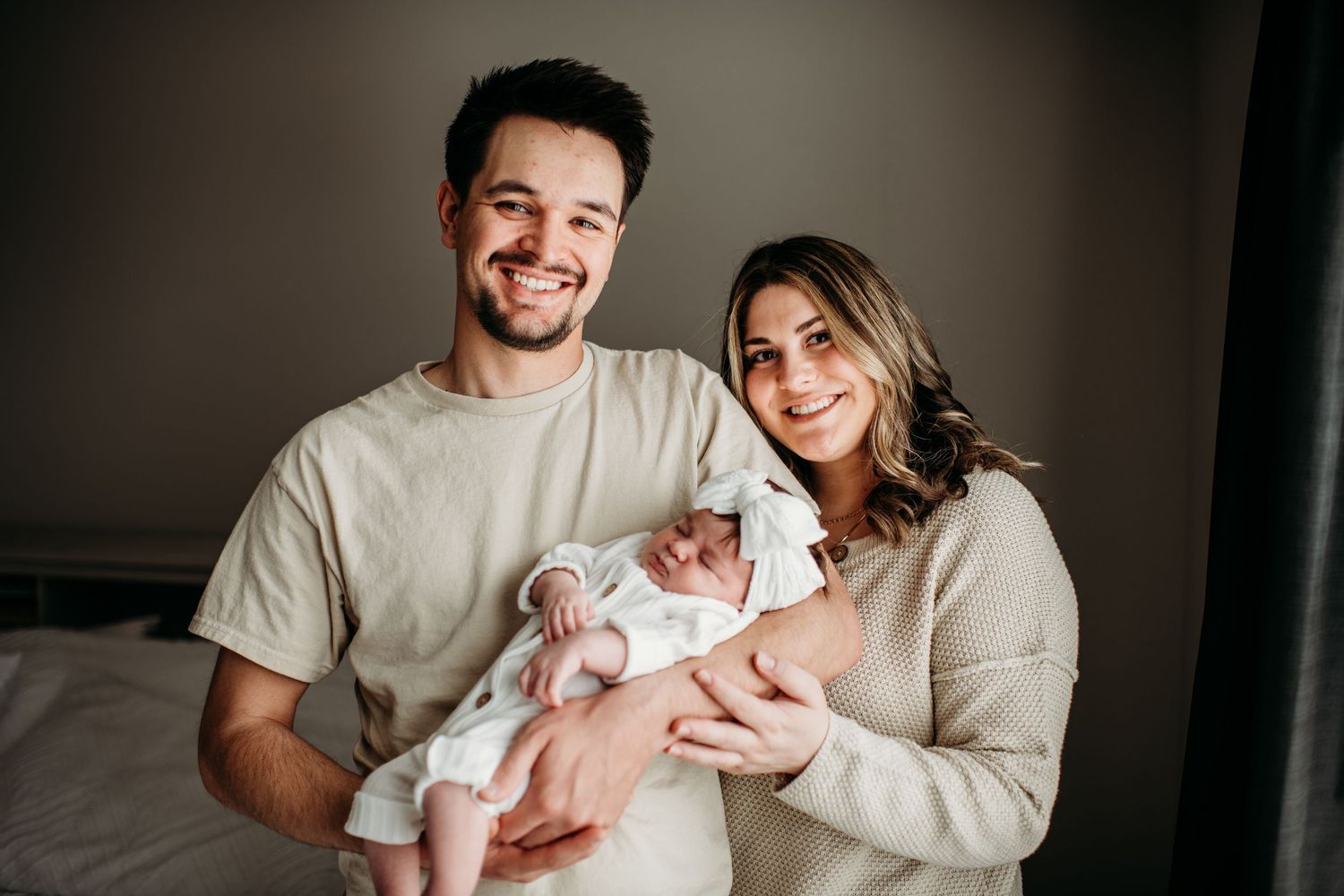Two parents in beige clothing hold their newborn baby while sitting together in a bedroom.