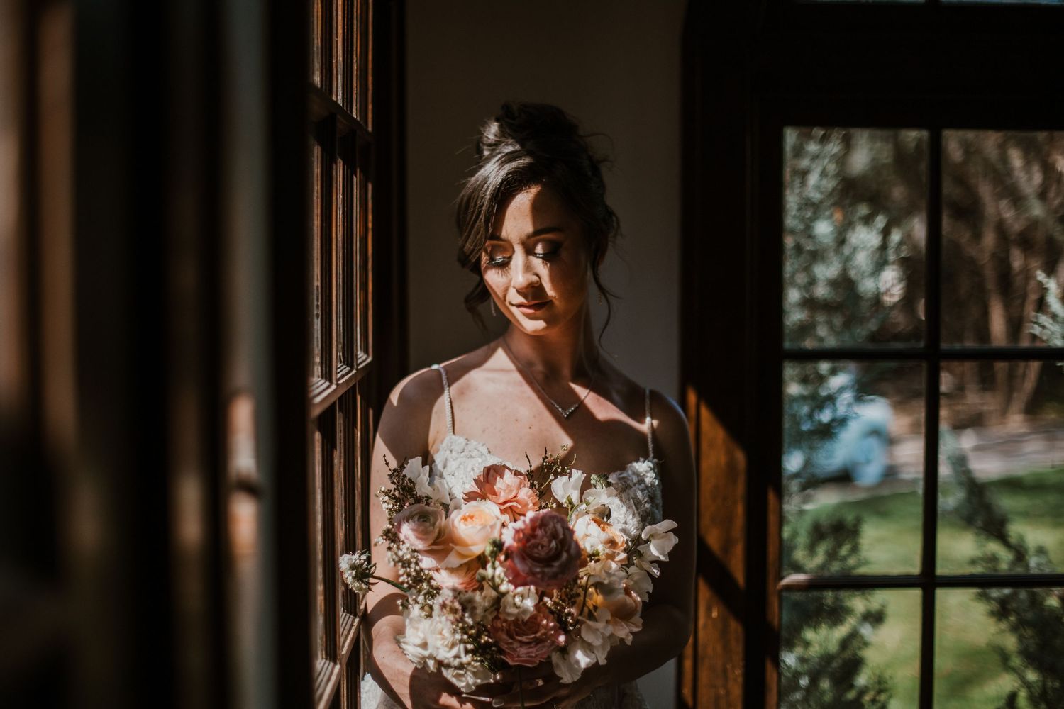 A figure in sunlit window holds a soft pink and peach floral bouquet while wearing bridal attire.