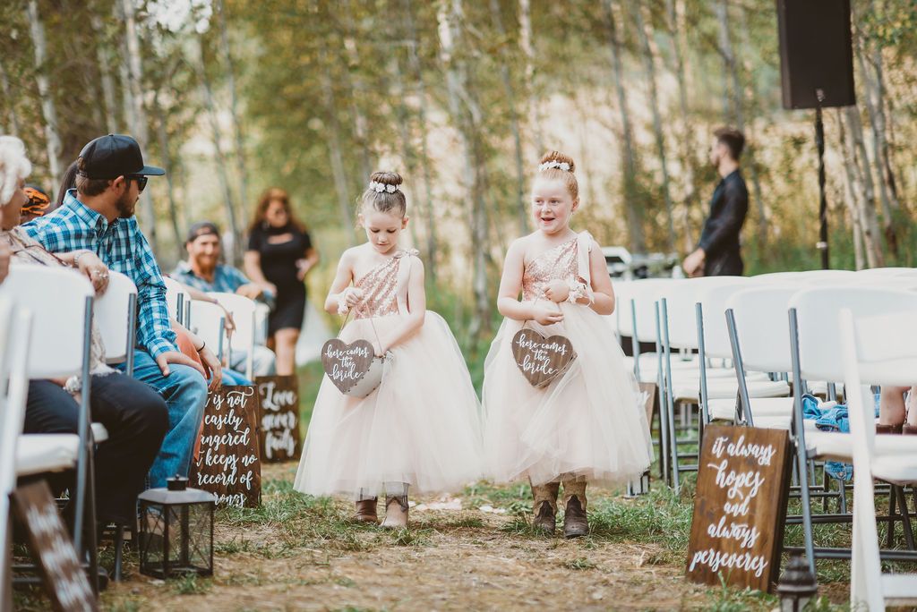 Two flower girls in pink tulle dresses hold heart-shaped signs during an outdoor forest wedding ceremony.