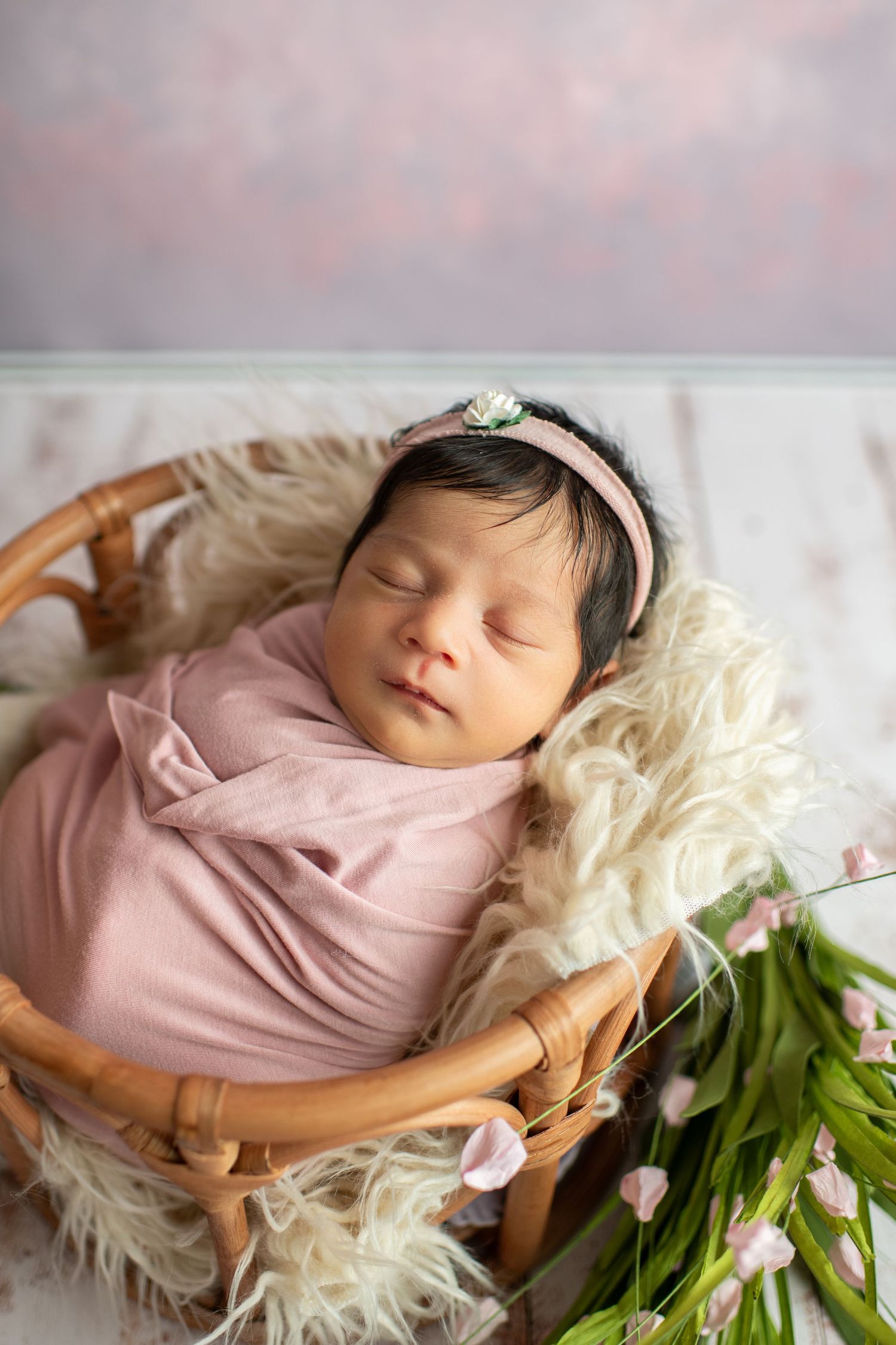 Newborn in pink swaddle and tiara rests in wooden basket with fur lining and white flowers.