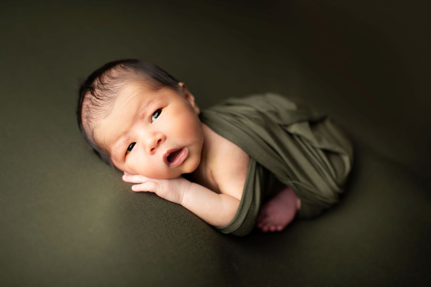 Newborn baby wrapped in olive green fabric on dark background in artistic portrait style.