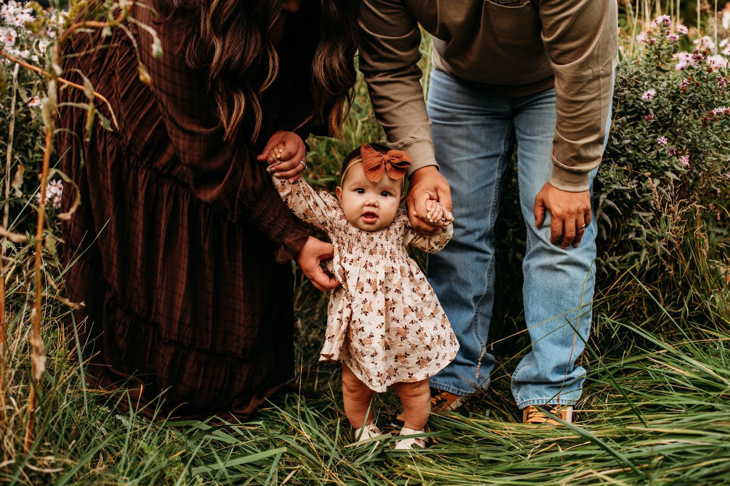 Little one taking first steps while holding parents' hands in garden setting.