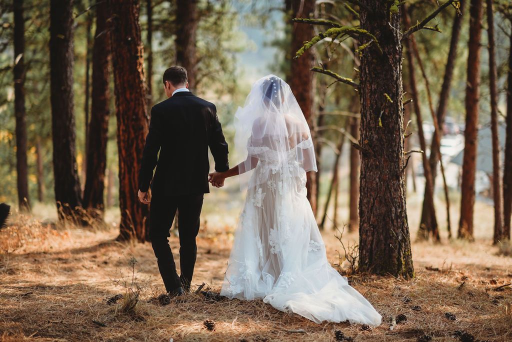 A couple walking hand in hand through a sunlit forest path in their wedding attire.
