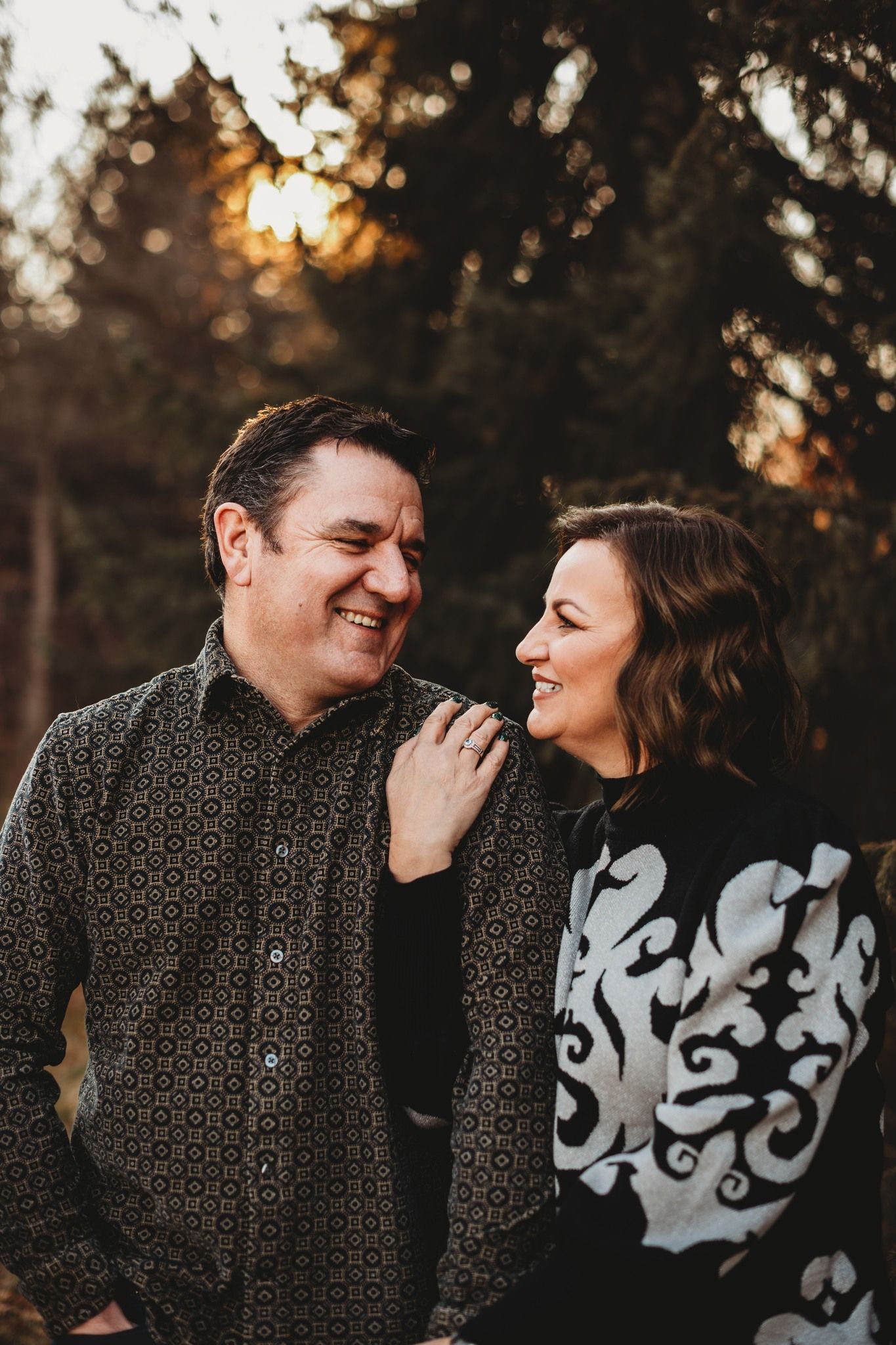 A couple shares a candid laughing moment at sunset among trees.
