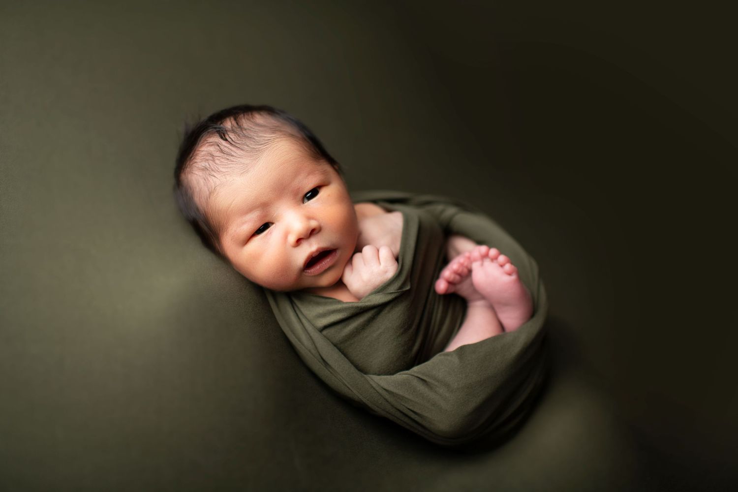 Newborn baby swaddled in olive green wrap against dark background.