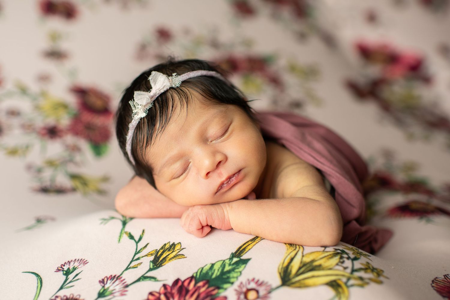 Sleeping newborn on floral backdrop wearing delicate white headband and swaddled in pink fabric.