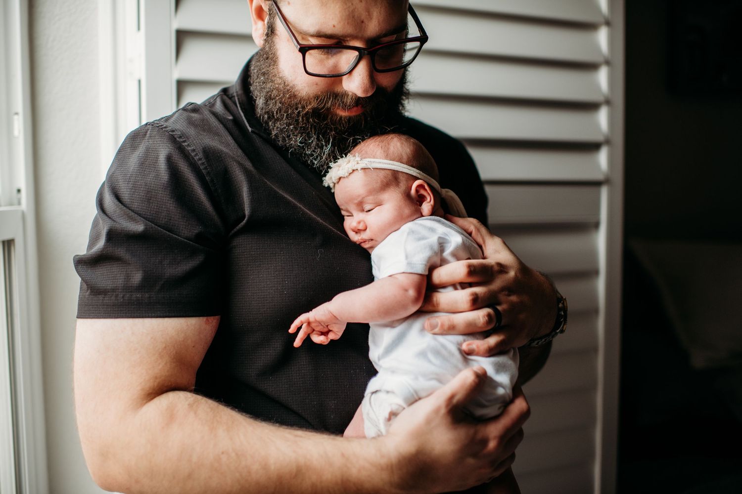 A bearded person wearing glasses and dark shirt tenderly holds a sleeping newborn baby against their chest.