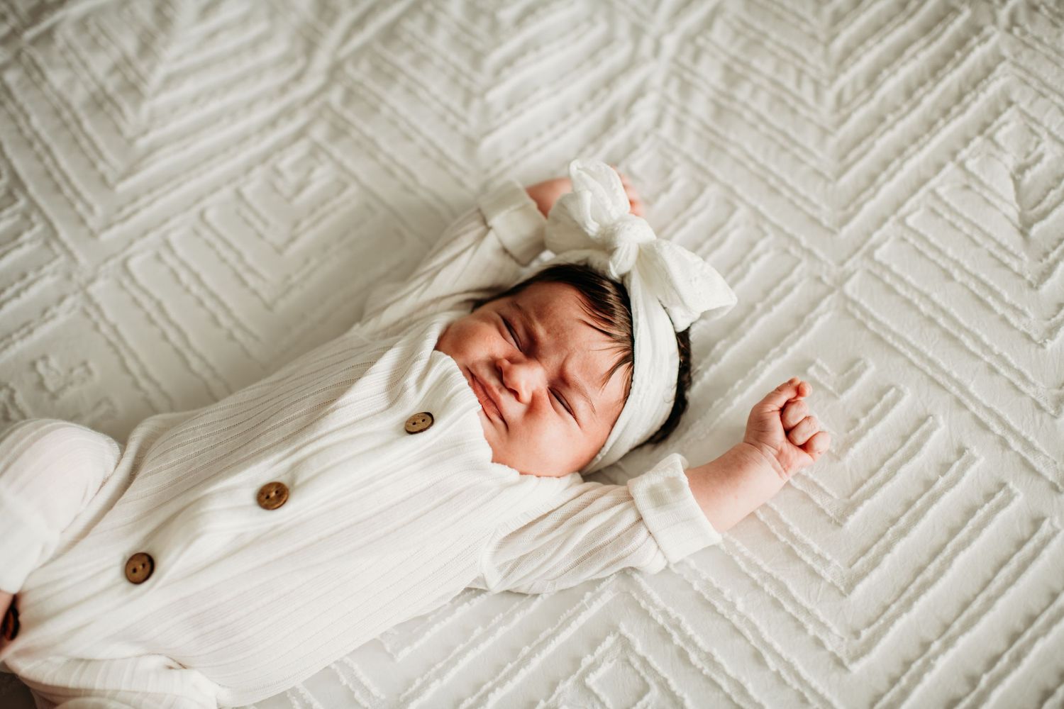 Baby wearing white hooded outfit smiles while lying on textured white blanket.