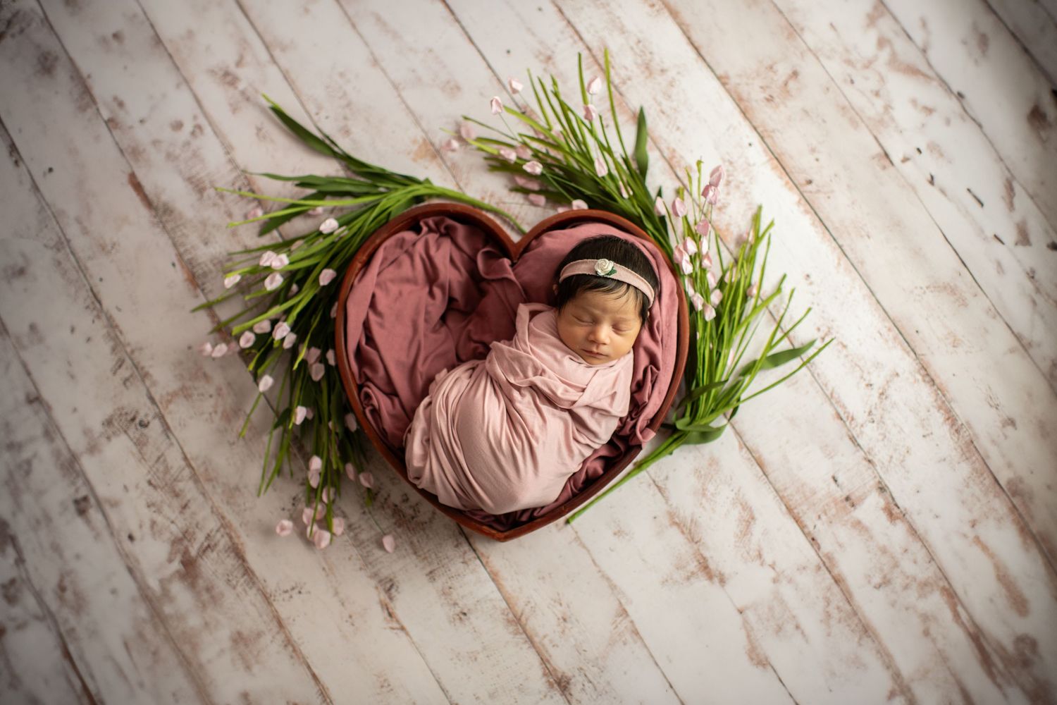 Heart-shaped basket with pink fabric and greenery on a whitewashed wood background in newborn photography setup.