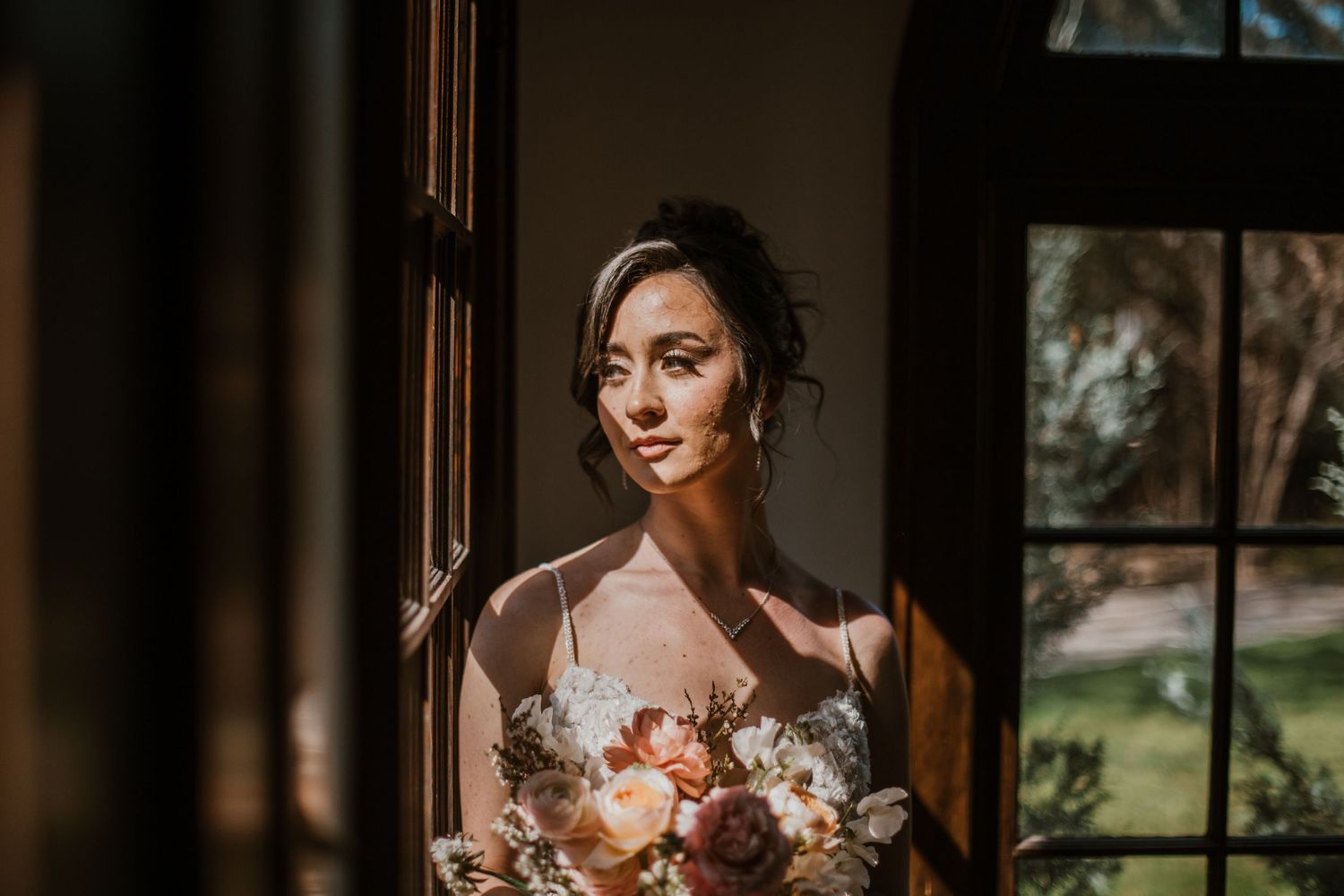 Sunlit figure at window holding beautiful peach and ivory wedding bouquet in moody interior setting.