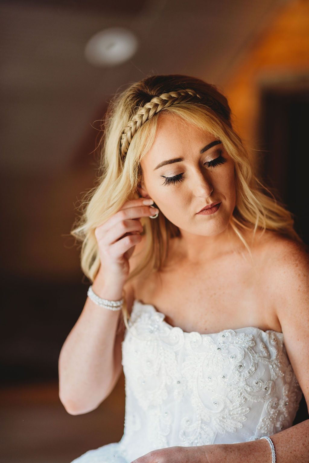 A bride wearing a white lace strapless wedding dress touches her earring while looking down with a braided hairstyle detail.