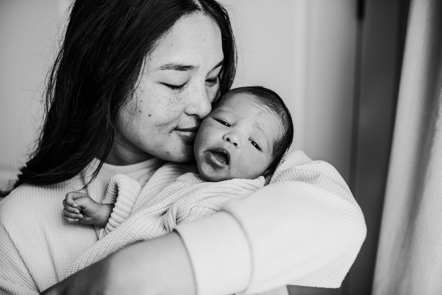 A tender black and white portrait shows a woman in a white sweater cradling and gazing lovingly at a yawning baby.