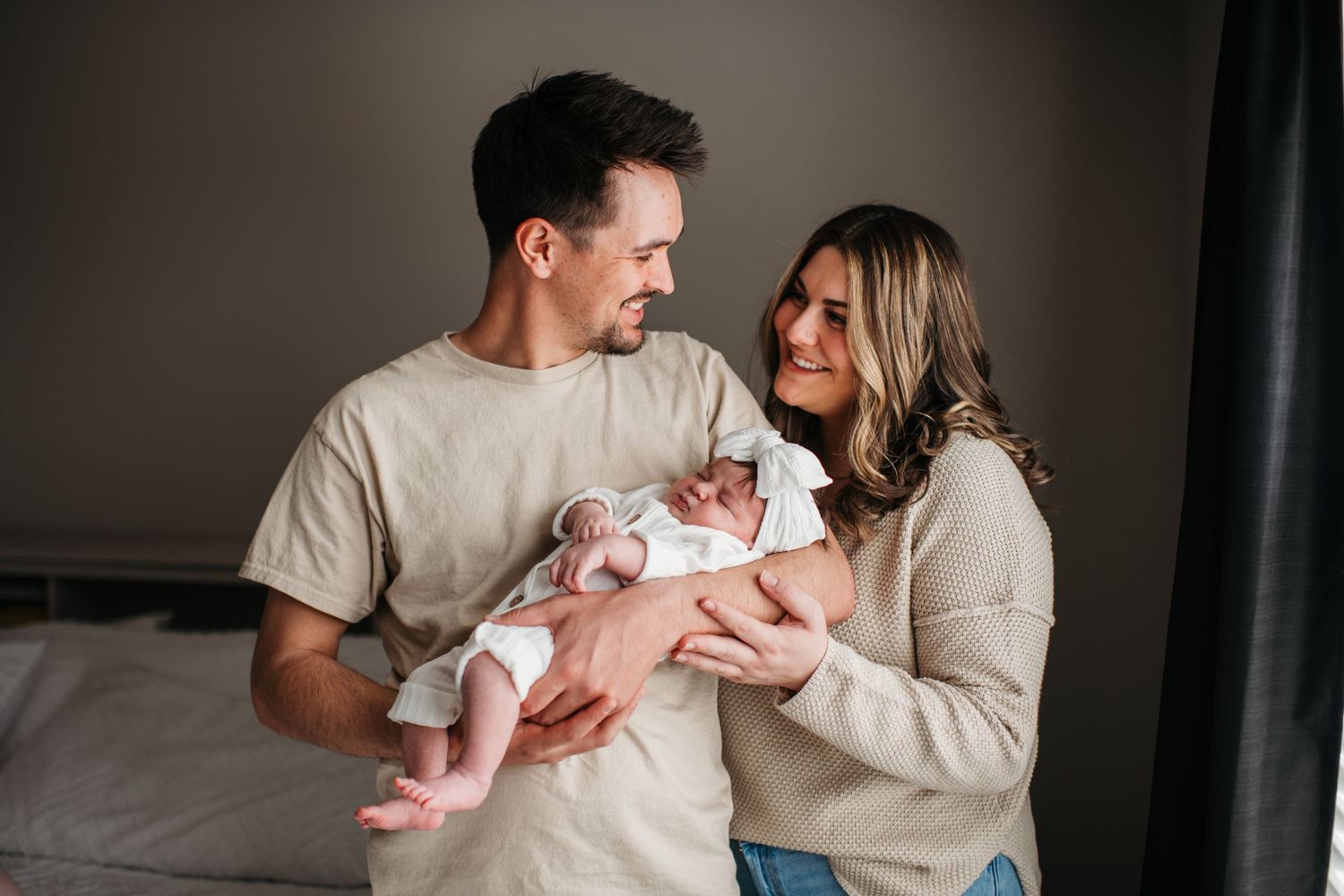 Couple in casual clothing tenderly holding newborn baby in bedroom during intimate family moment.