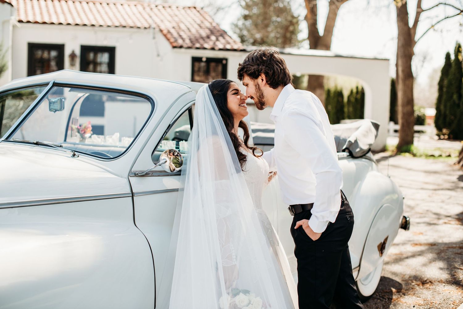 A couple shares a tender moment beside a vintage light gray car during their wedding day celebration.