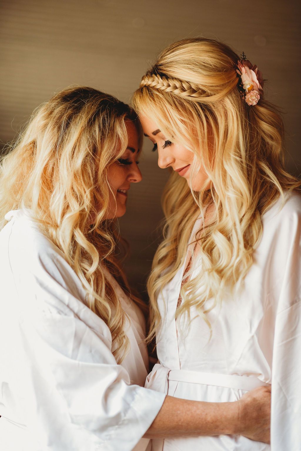 Two women in white robes share an intimate pre-wedding moment with matching blonde wavy hairstyles and braided details.